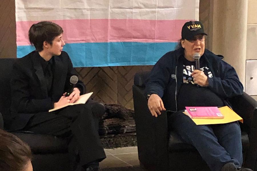 Two people sit in front of the transgender pride flag and address an audience.